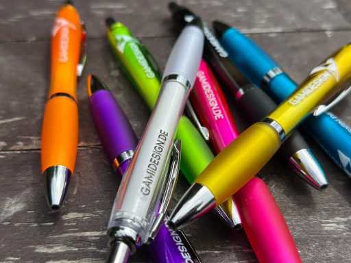Bunte Kugelschreiber (orange, lila, blau, pink, grün, silber) auf einer Holzoberfläche.