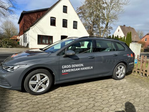 Grauer Kombi mit Beschriftung an einer Straße vor einem Wohnhaus.