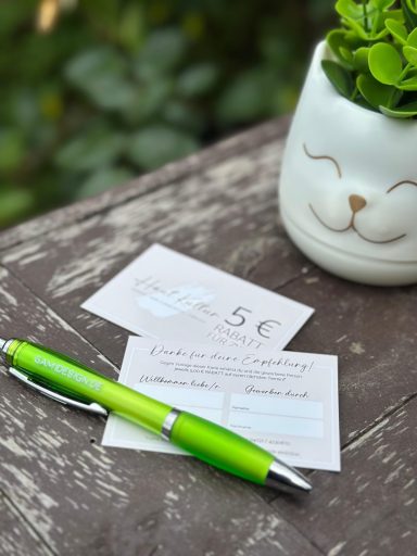 Grüner Stift liegt auf einer Holzoberfläche neben zwei Gutscheinen und einem Pflanzenübertopf.