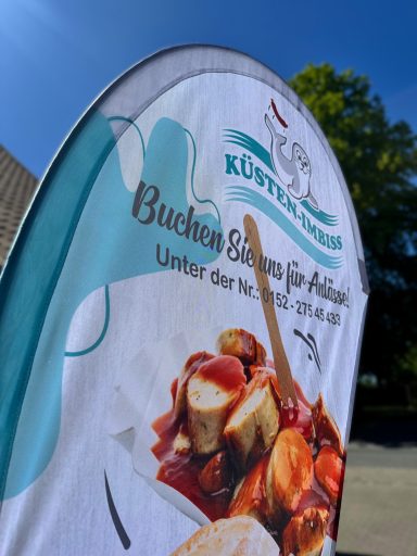 Werbeplakat für Bratwürste mit Slogan und bunten Zutaten, vor blauem Himmel.