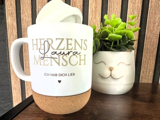 Eine Tasse mit der Aufschrift "Herzensmensch" neben einem lächelnden Pflanzentopf.
