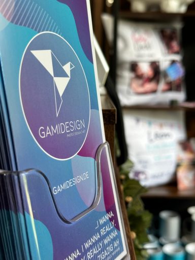Bunte Broschüre mit dem Titel „GAMIDESIGN“ in einem Informationsständer.